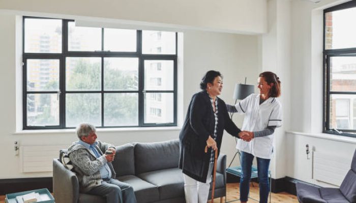 912081976-612×612 Friendly female nurse assisting senior woman to walk with cane in nursing home and man sitting on sofa having coffee